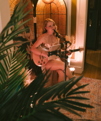 girl seated with guitar, singing into mic, plant in front of her