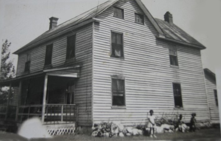 A black and white image of a two-story wooden residence featuring a porch; two children and several goats are seen near its side.