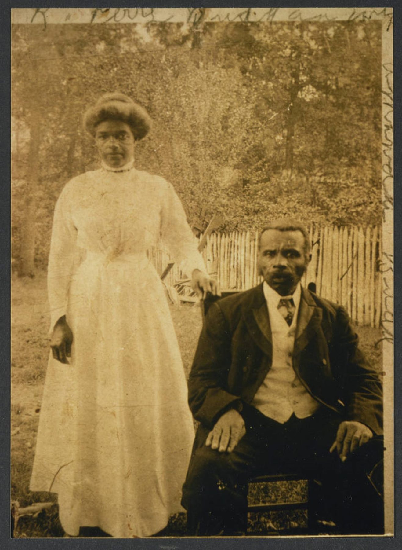 A woman poses next to a seated man outdoors, both dressed in early 20th-century formal attire, with a wooden fence and trees in the background.