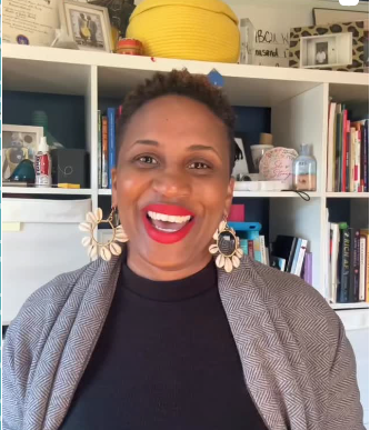 woman with short dark hair smiling at camera wearing black top and gray sweater and large ivory hoop earrings