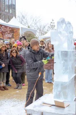 man in hat and brown pants and jacket holding a saw carving large block of ice into a sculpture while a crowd behind him watches