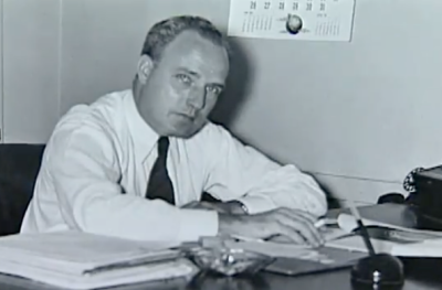 B&w photo of a man in white shirt and dark tie sitting at a desk