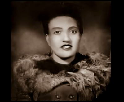 B&w photo of a woman looking at the camera, adorned in a fur-trimmed coat