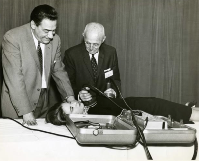 B&w photo of two men in suits using a defibrillator on a woman lying on a table
