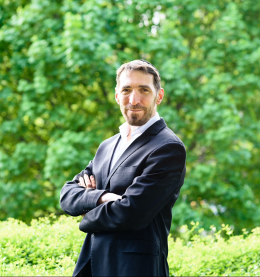 man in dark blazer and white shirt standing with arms crossed and facing camera with closed-mouth smile, standing in front of trees