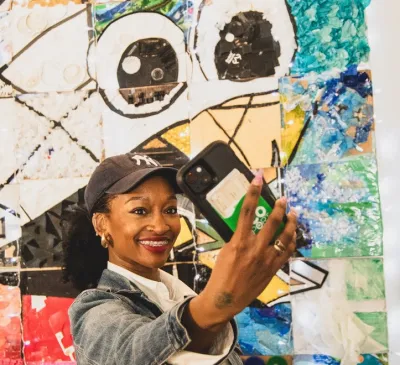 A woman in a baseball cap takes a selfie in front of Trash Wheel art