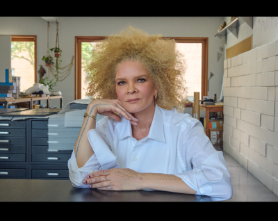 woman in blue blouse and blonde afro sits at table looking at camera with one hand under chin