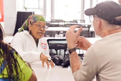 two people at a table having a conversation, one in white sweater, glasses and head scarf with hand on table, the other with back to camera wearing brown baseball cap and tan sweater with elbows on table and hands together.