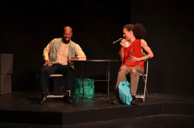 Two people sitting in chairs on stage, both smiling, looking at each other, person on right holding microphone speaking to person on left