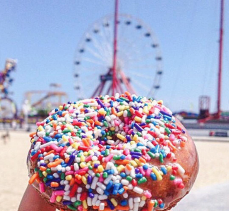 close up of rainbow sprinkles on a donut with beach and ferris wheel in background