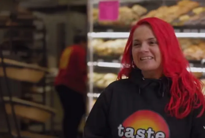 A woman with bright cherry red hair stands in a grocery store. She wears a black hoodie with the word "taste" in all lower-case letters against a red, orange, and yellow gradient.